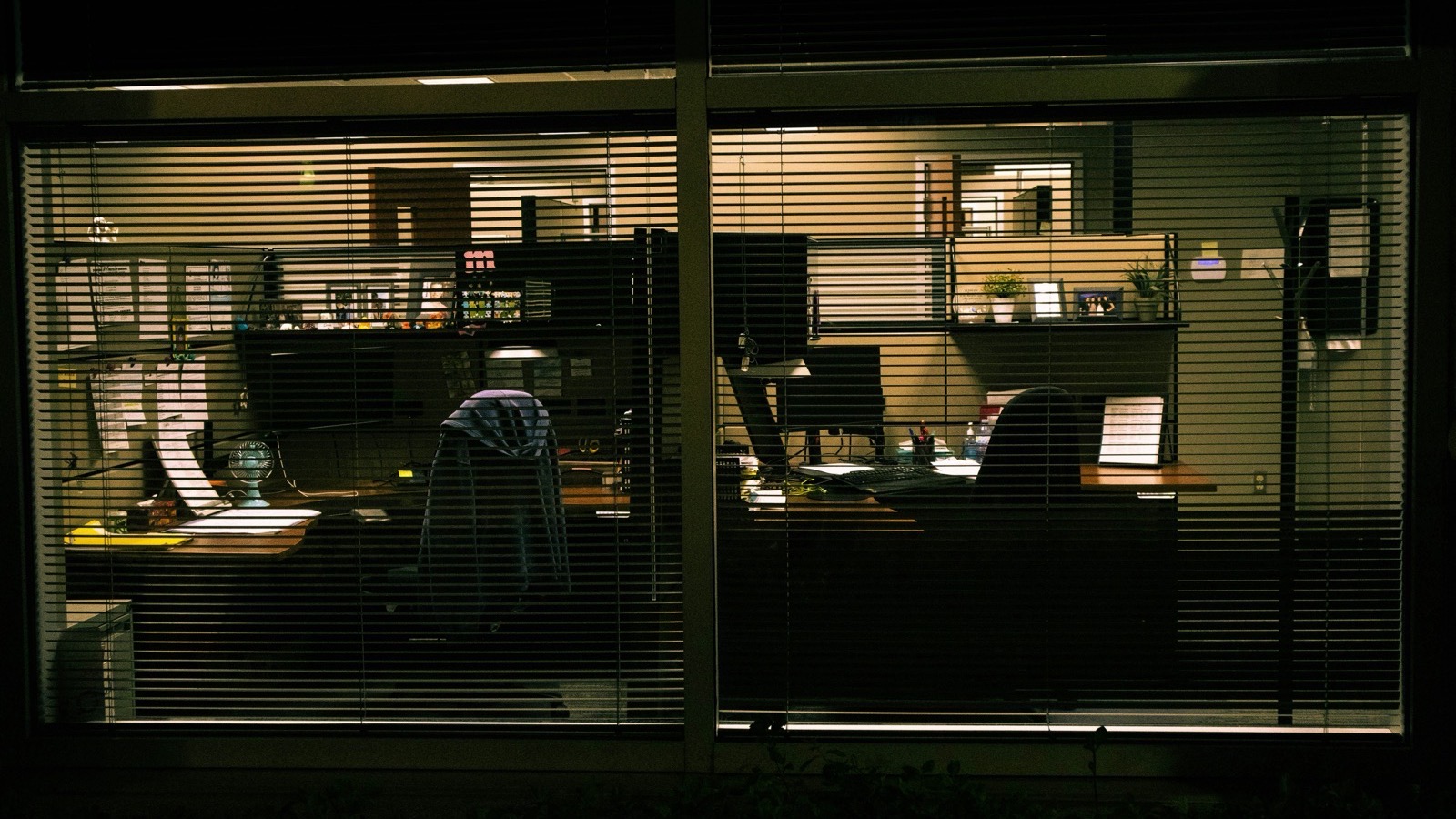 Empty office at night seen through window blinds, screens still glowing while systems keep running
